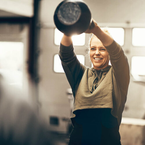 Fit young woman enjoying an exercise class at the gym Fit young woman in sportswear smiling while swinging a dumbbell over her head during a workout session in a gym
